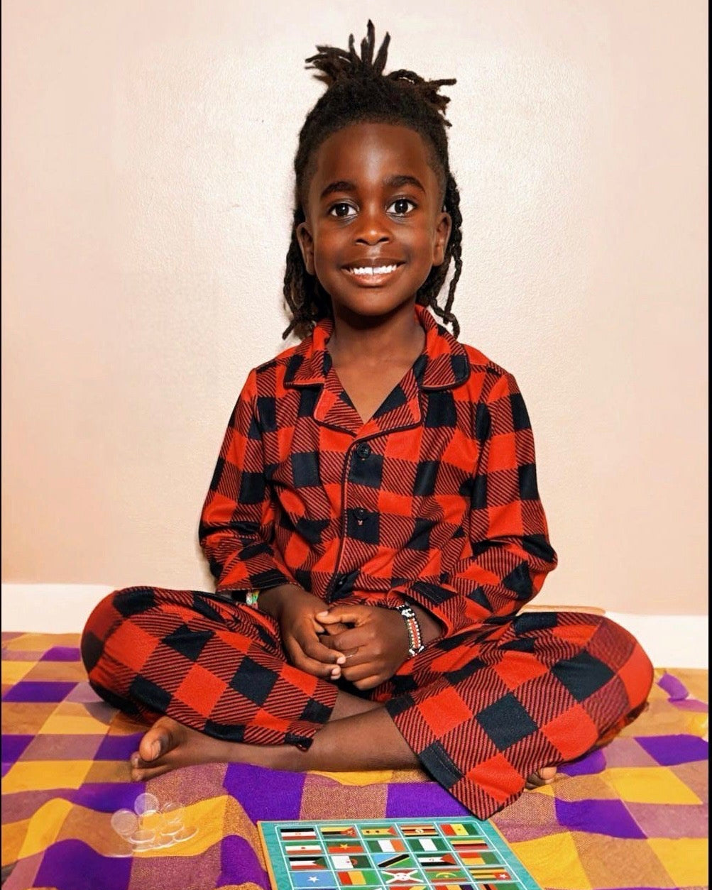 Child wearing red and black checkered pajamas sitting on a colorful checkered rug.